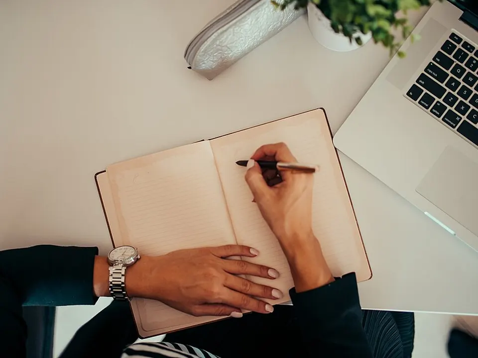 Person writing in a notebook while using a laptop at a workspace